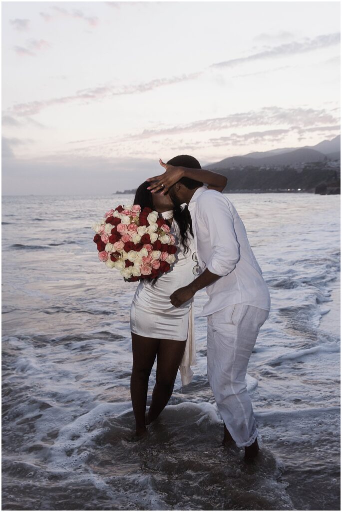 Couple kissing with feet in the water and holding a floral bouquet at Will Rogers State Beach, Malibu