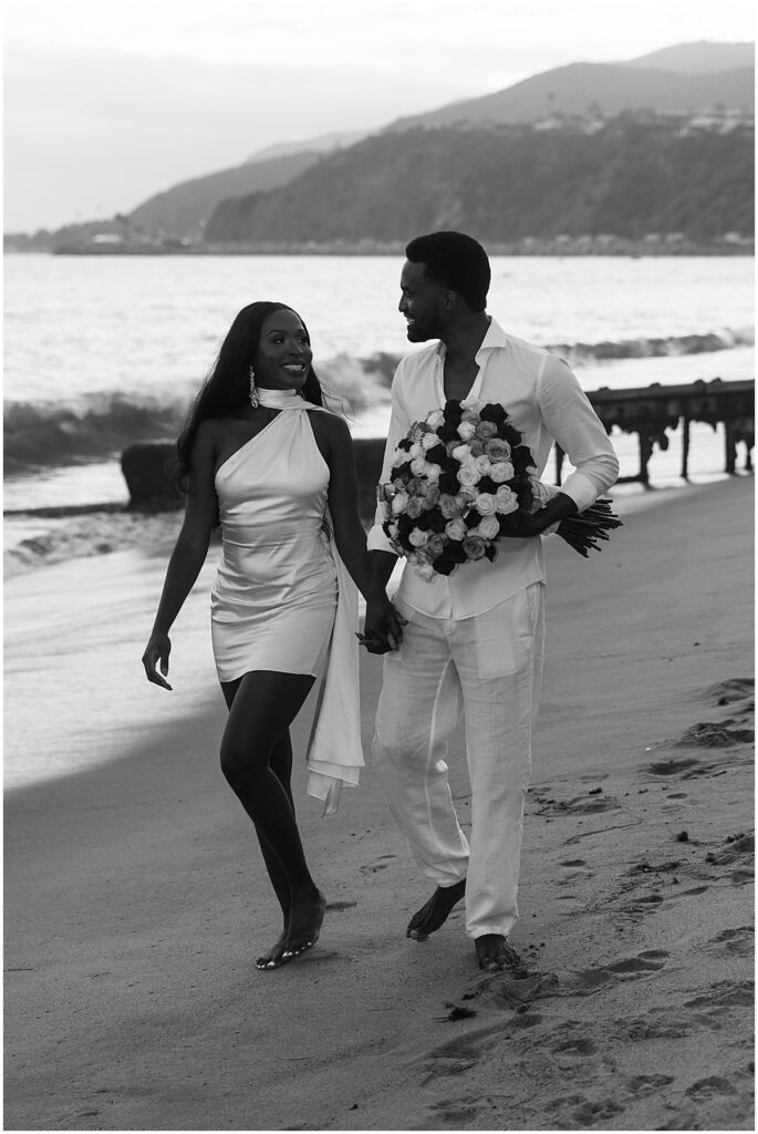 Couple walking along beach carrying large bouquet of flowers