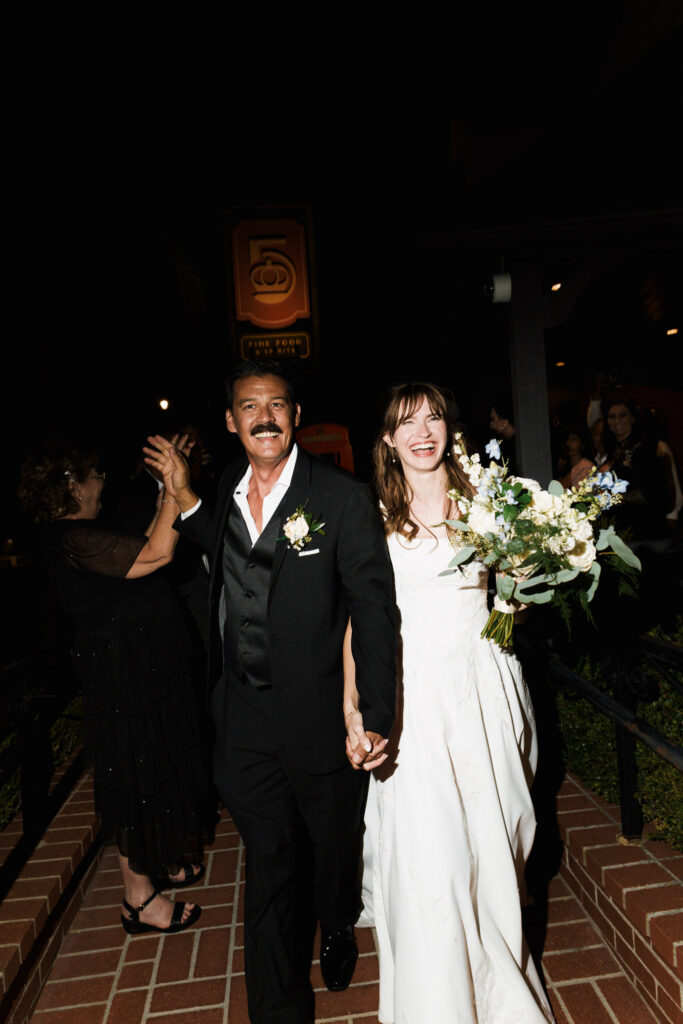 Bride and groom walking out at night as guests celebrate, bouquet in hand
