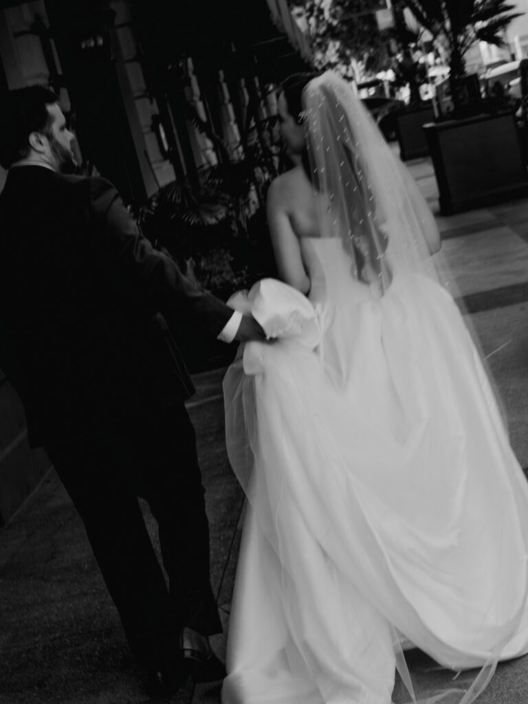 Motion-filled black and white portrait from behind as the newlyweds walk down the city sidewalk, her pearl-studded veil trailing in the breeze.