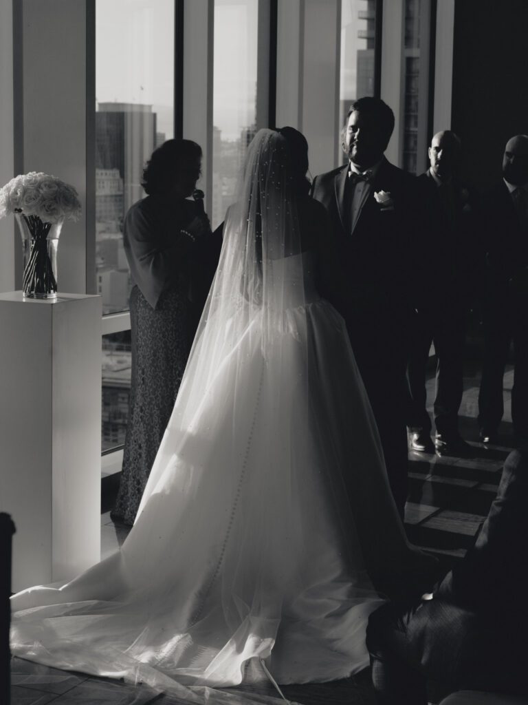 Moody black and white capture of the bride and groom exchanging vows in front of high-rise city window panes and white rose pedestals .