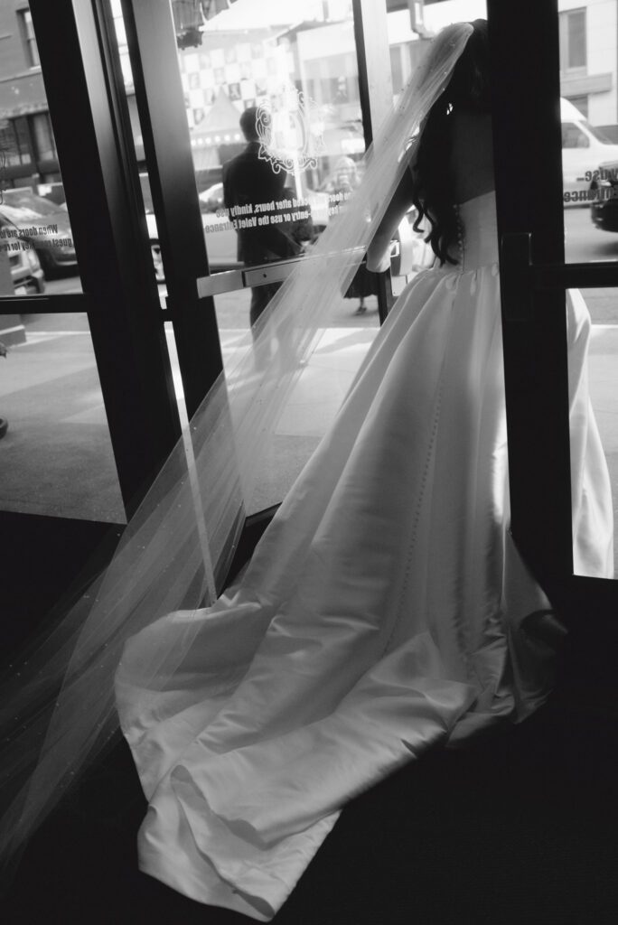 Back view of the bride with flowing pearl veil and long train pushing open glass hotel doors toward the groom waiting outside on the street.