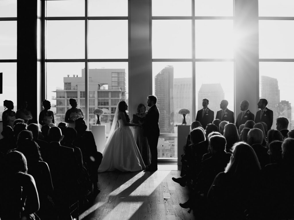 Black and white wide shot of wedding guests seated in silhouette watching the bride and groom exchange vows in front of sunlit high-rise windows .