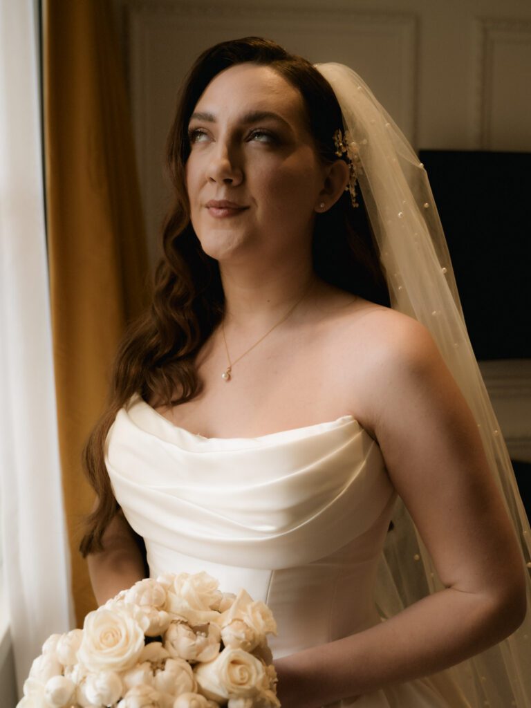 Close-up bridal portrait of the bride wearing an off-the-shoulder cowl neckline dress, gold necklace, and a delicate pearl-studded veil in soft window light.