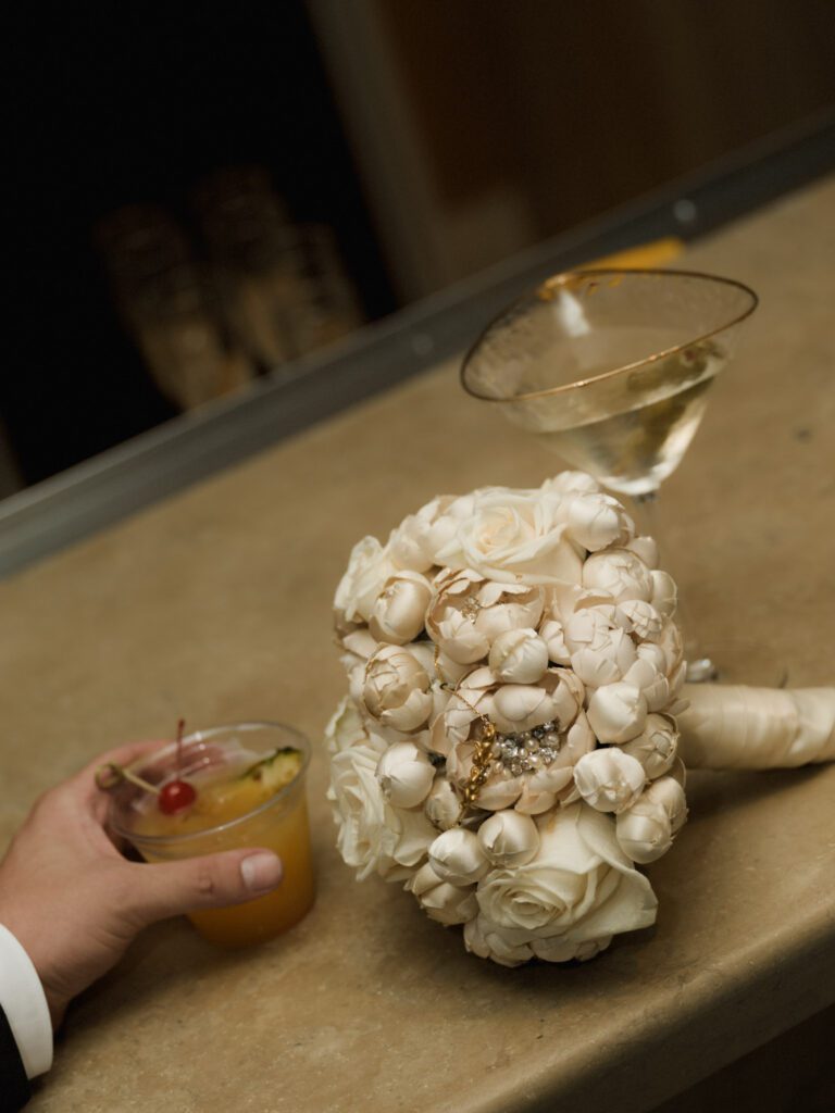 Textured white brooch bridal bouquet resting on a sleek bar counter next to an olive martini and a tropical garnished cocktail .