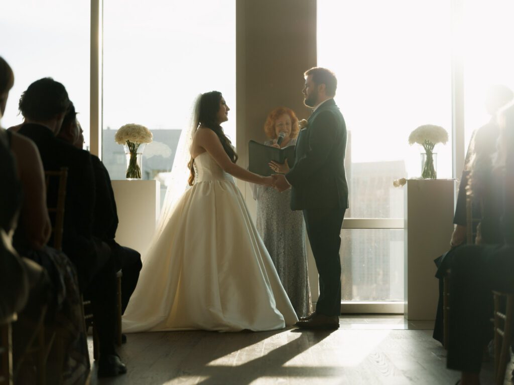 Bride and groom holding hands and smiling at each other during their ceremony vows with the officiant standing between them in bright window light .