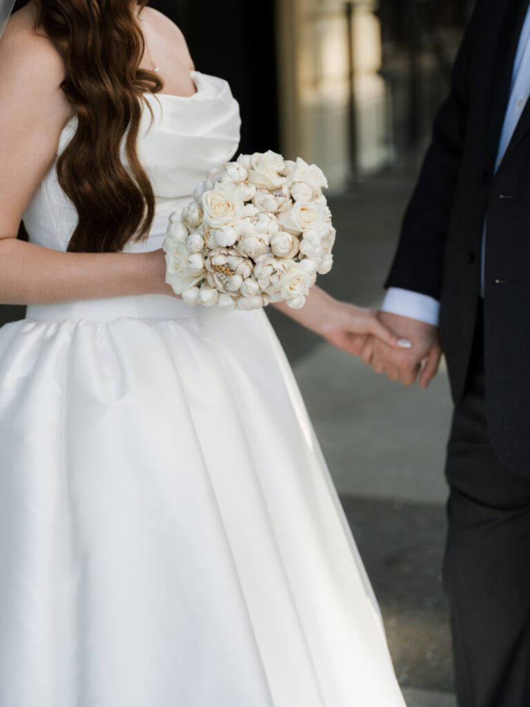 Close-up of the newlyweds holding hands on the sidewalk, showcasing the bride’s textured white rose bouquet and classic satin wedding dress.