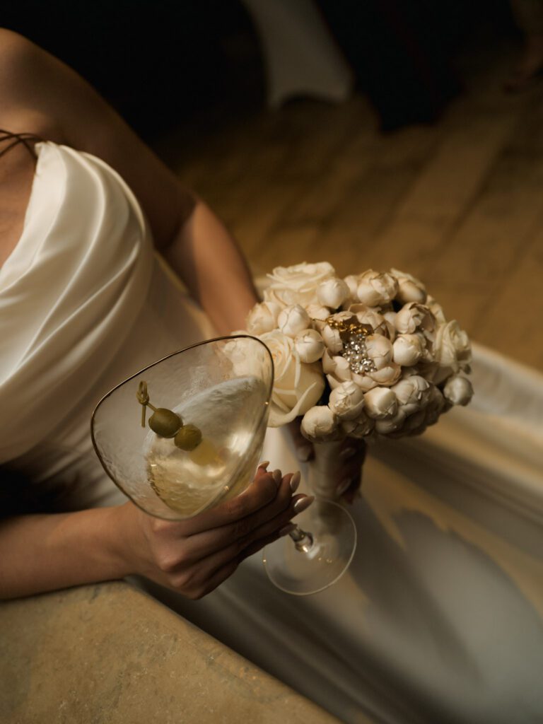 Overhead perspective of the bride holding an olive-garnished dirty martini glass in one hand and her textured white rose bouquet in the other .