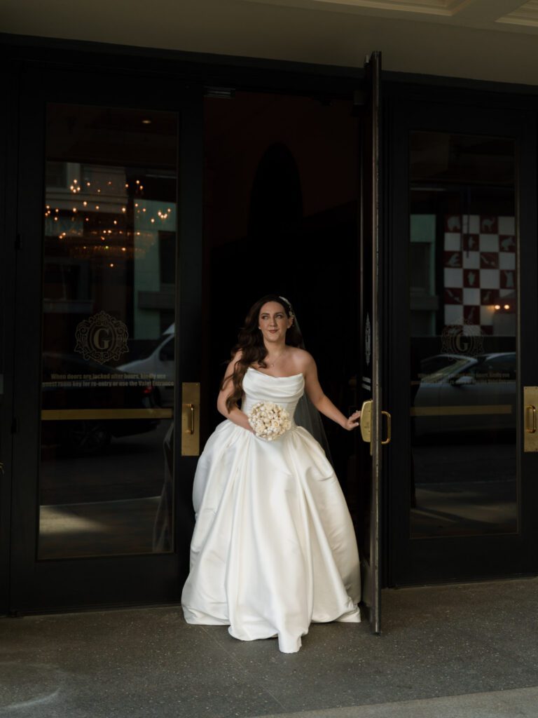 Bride in full satin ballgown holding her bouquet as she opens the exterior glass hotel doors to step out onto the city sidewalk.