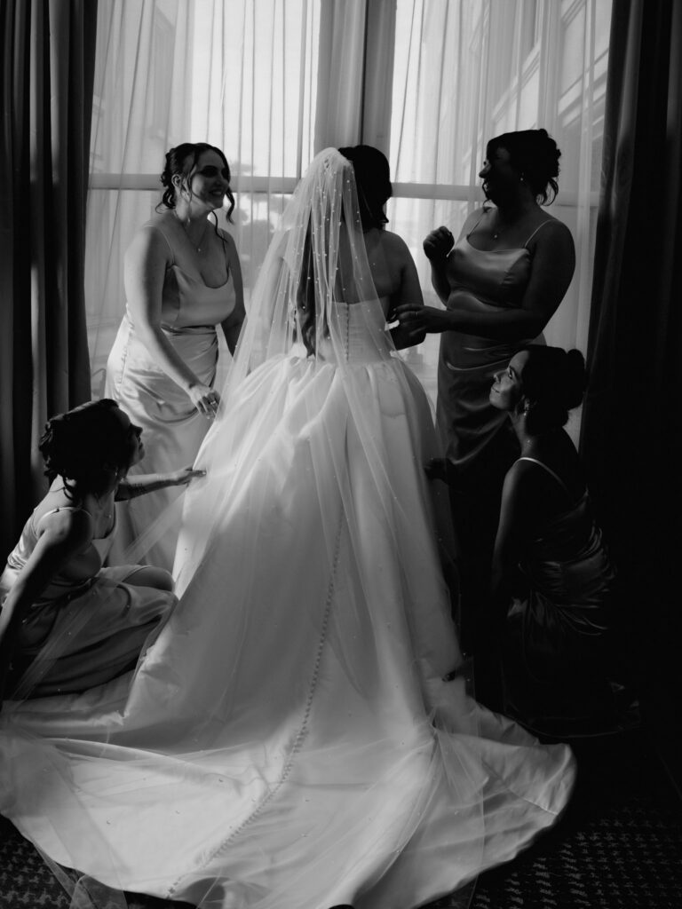 Black and white view of the bride from behind wearing a pearl-studded cathedral veil while four bridesmaids adjust her gown in front of tall windows.