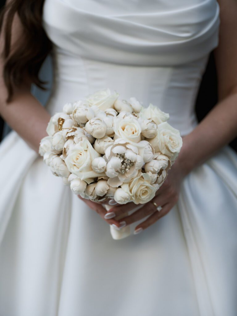 Detailed close-up of the bride's hands holding her white silk floral bouquet, showing her gold wedding band and jeweled brooch details tucked into the blooms.