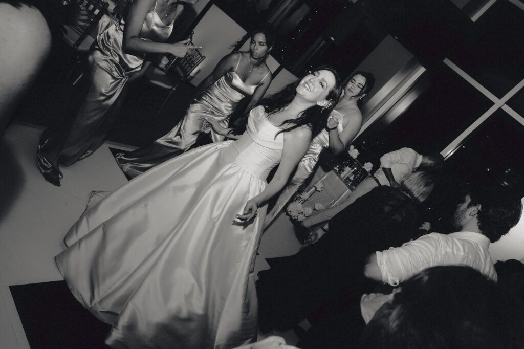 Dynamic black and white wide-angle photograph of the bride dancing joyfully in her ballgown amidst a crowded wedding dance floor .