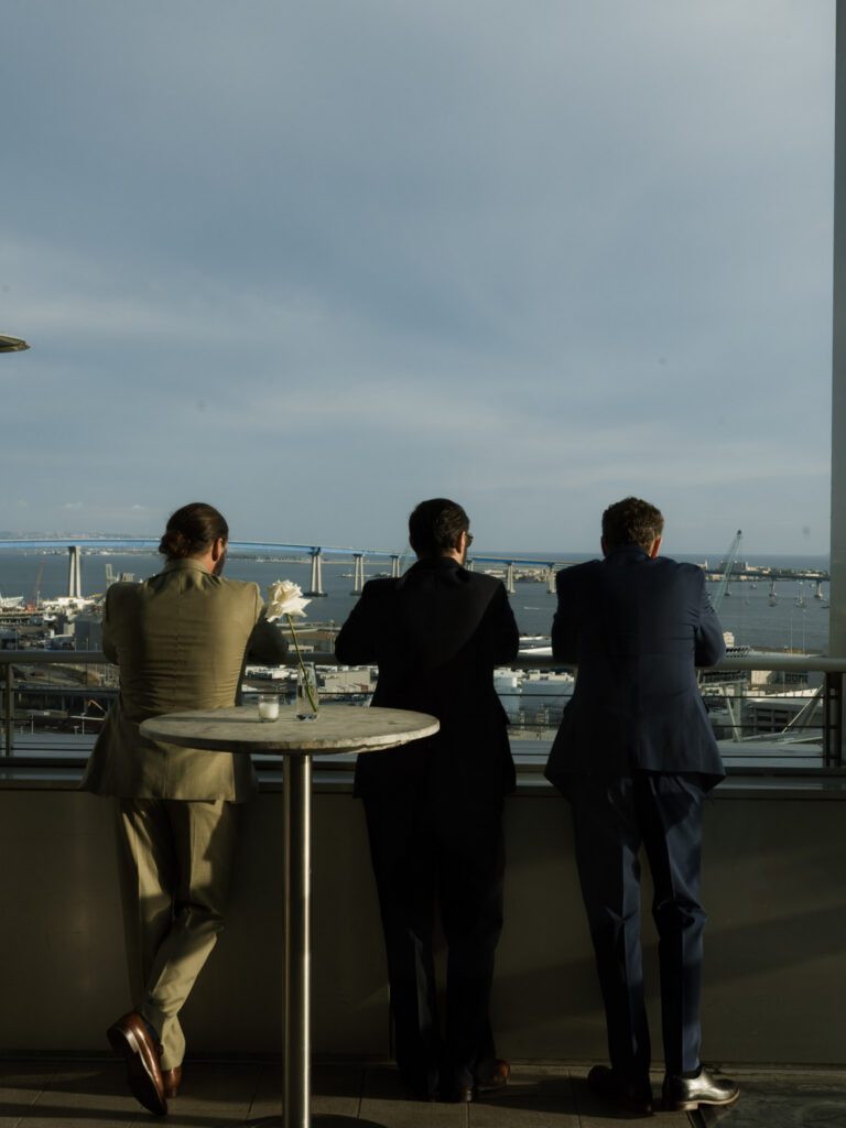 Three suited male guests leaning against the terrace glass railing taking in the panoramic view of the Coronado Bridge and bay .