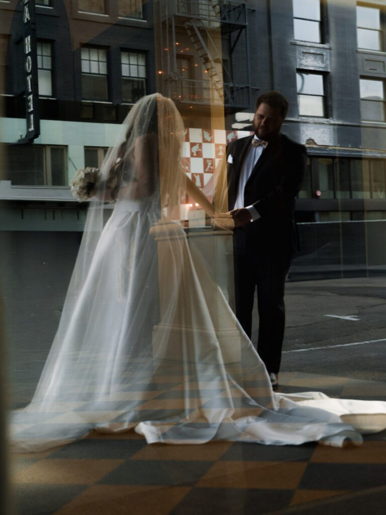 Creative reflection portrait through hotel window glass blending the bride and groom holding hands with the historic San Diego city streetscape.