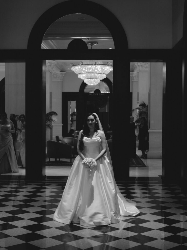 Symmetrical black and white photograph of the bride standing beneath a dramatic architectural arch and glowing chandelier in a historic hotel lobby.