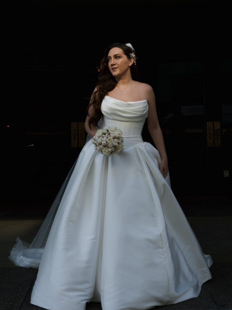 Bride in a strapless cowl-neck ballgown holding her bouquet while glancing softly to the side in front of dark entryway hotel doors.