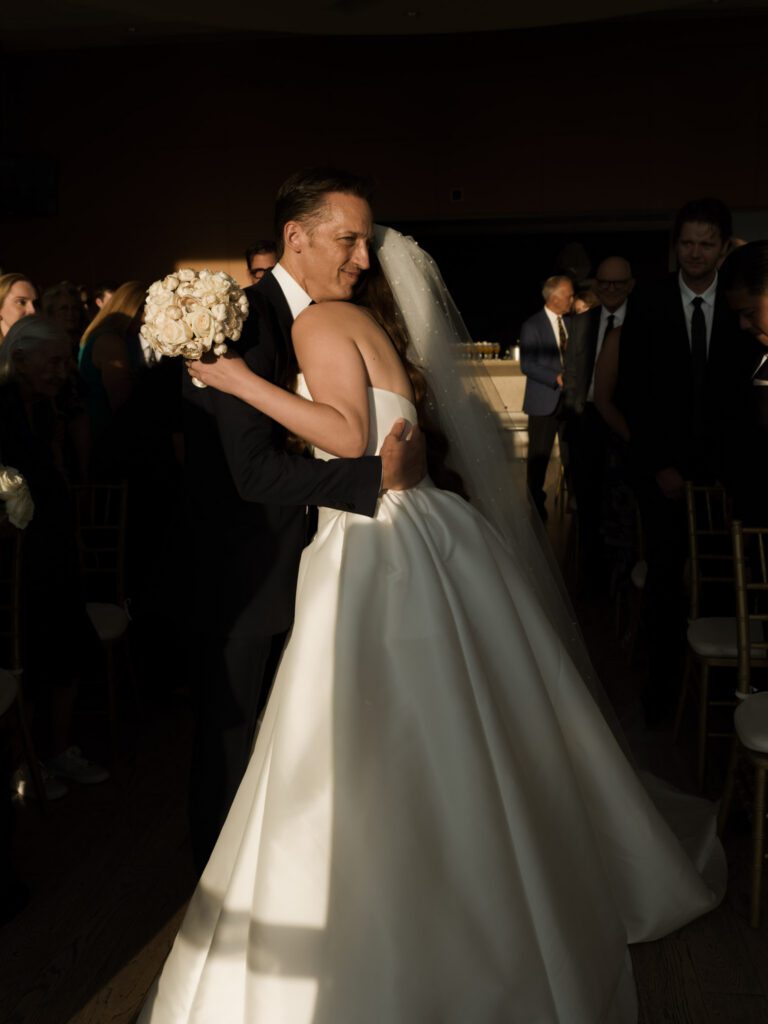 Emotional father of the bride smiling warmly as he hugs his daughter in her satin ballgown and pearl veil at the end of the ceremony aisle .