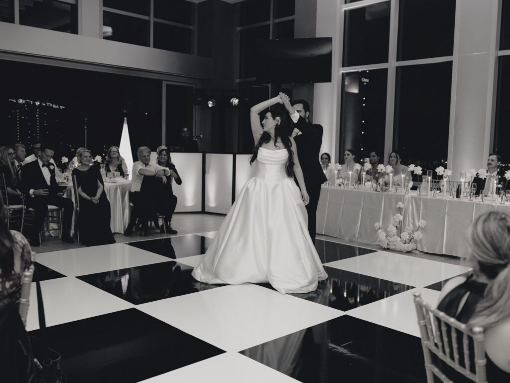Black and white capture of the groom twirling the bride in her ballgown across a glossy black and white checkered dance floor as wedding guests watch .