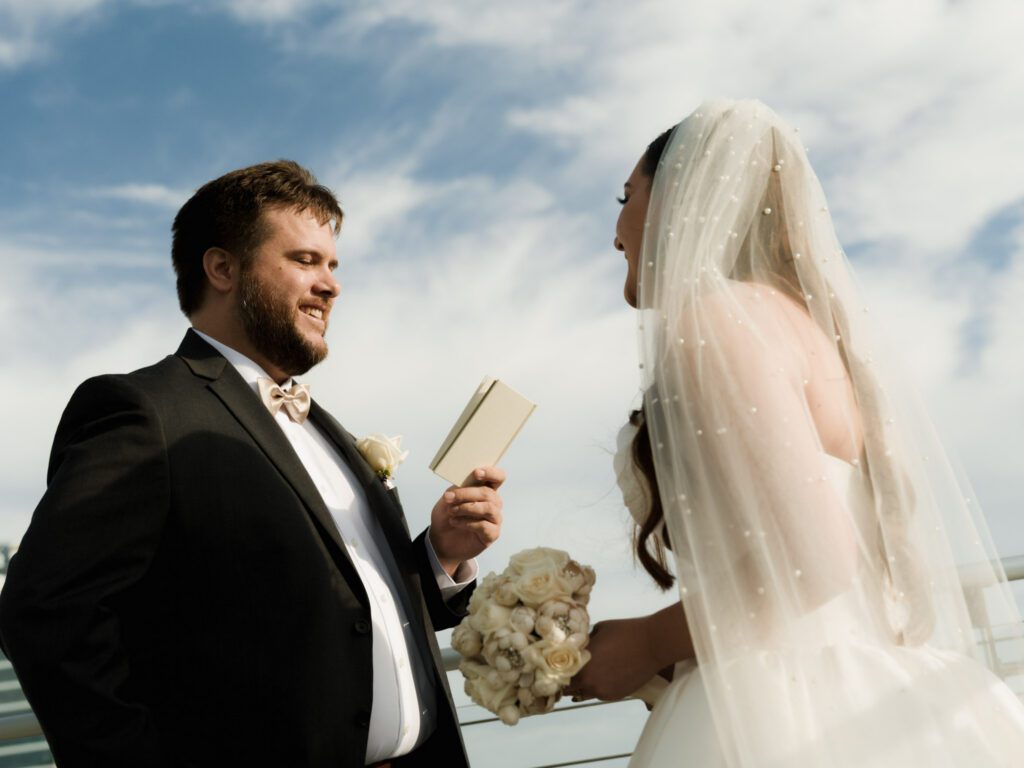 Groom smiling as he reads from a vow booklet to his bride in her pearl-accented veil under open blue skies on a high-rise terrace.