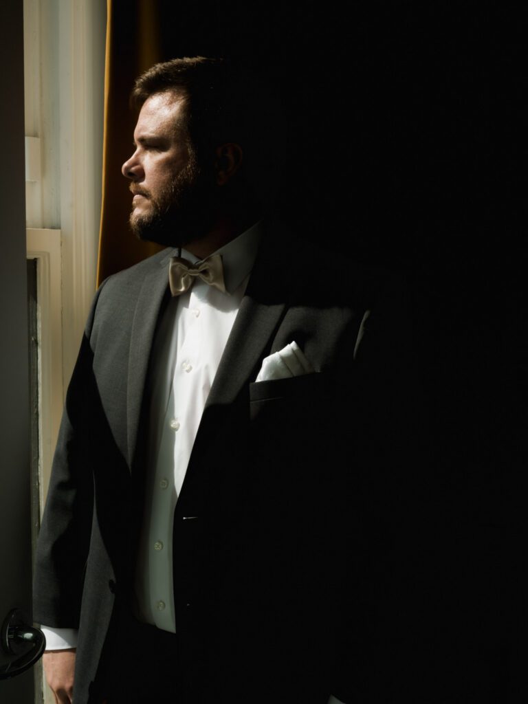 Moody editorial portrait of a bearded groom in a dark charcoal suit, champagne gold bowtie, and pocket square looking thoughtfully out a window.