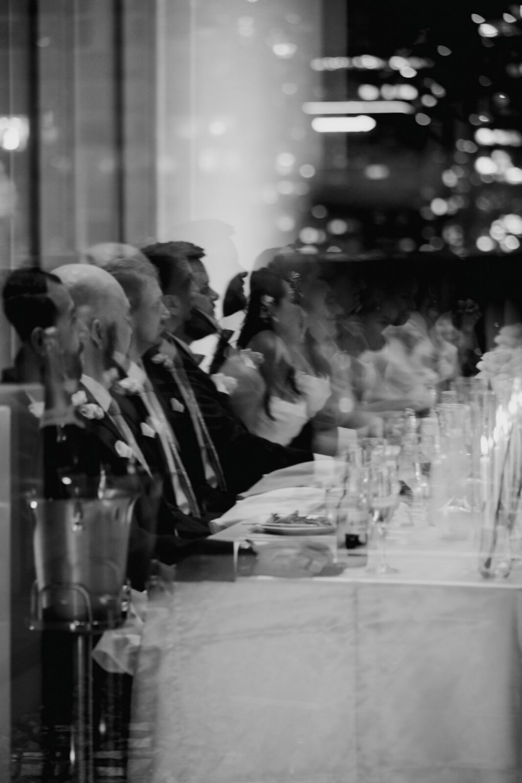 Dreamy black and white reflection through glass of the wedding party and newlyweds seated along a candlelit head table listening to speeches .