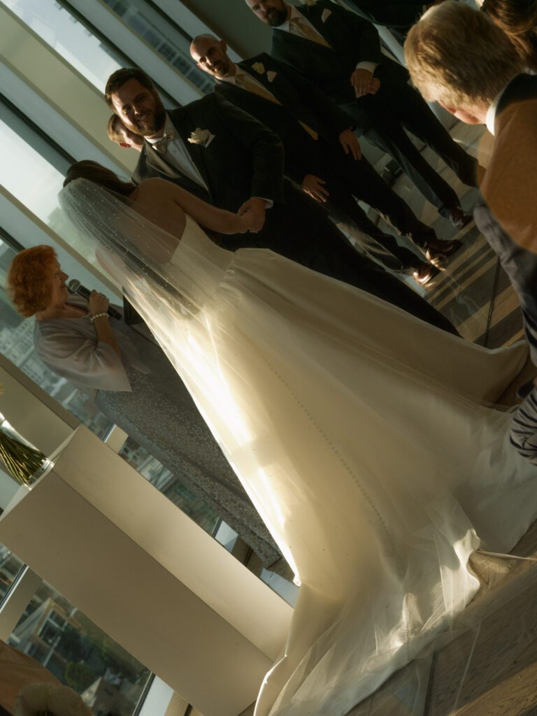 Elevated diagonal view of the bride in a satin ballgown with cascading pearl veil holding hands with the smiling groom during their vows .