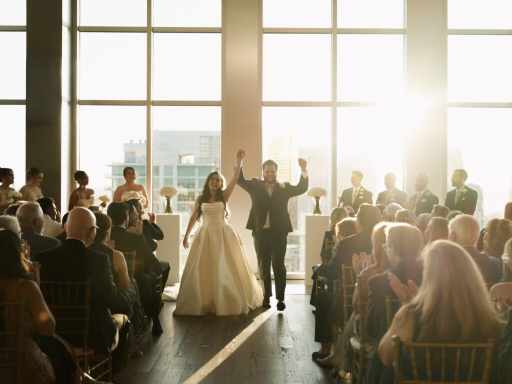 Newlyweds walking down the ceremony aisle with hands raised in celebration as seated wedding guests clap and cheer .