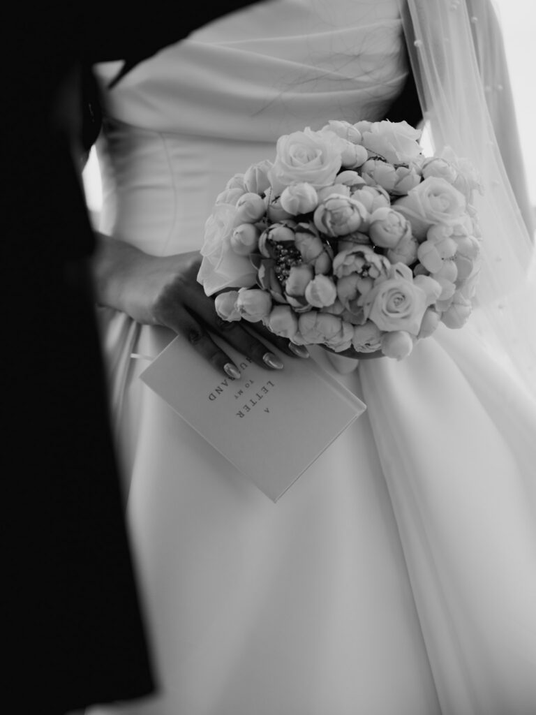 Black and white close-up of the bride holding a structured rose brooch bouquet alongside a vow booklet titled "A Letter to My Husband."