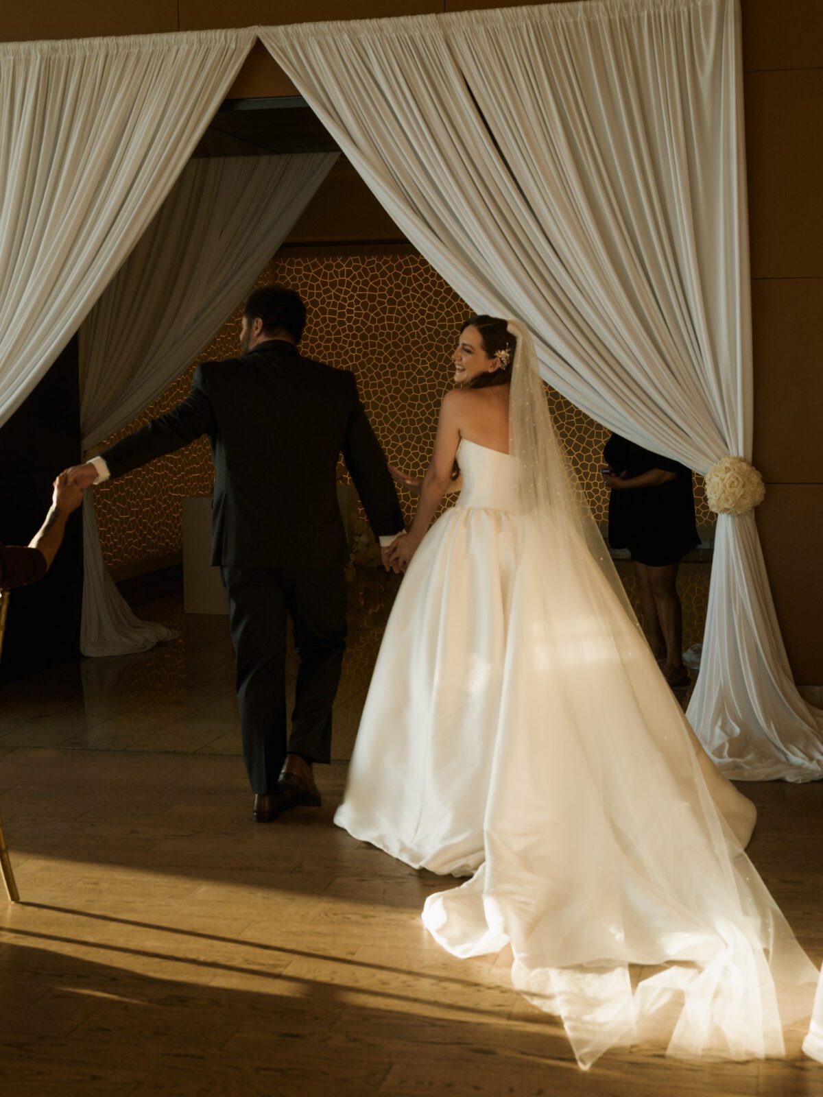 Back view of the bride and groom holding hands and smiling as they walk through white draped curtains, showing off the full satin gown train .