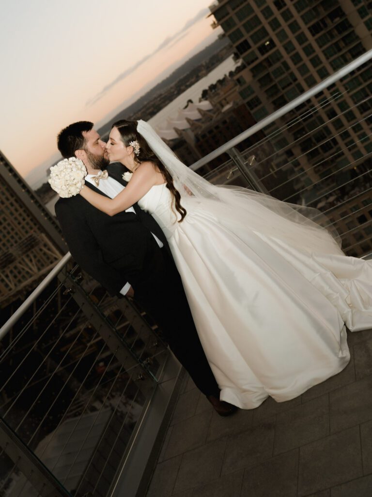 High-angle shot of the bride in a satin ballgown and groom kissing by a glass balcony railing with downtown city high-rises below at dusk .