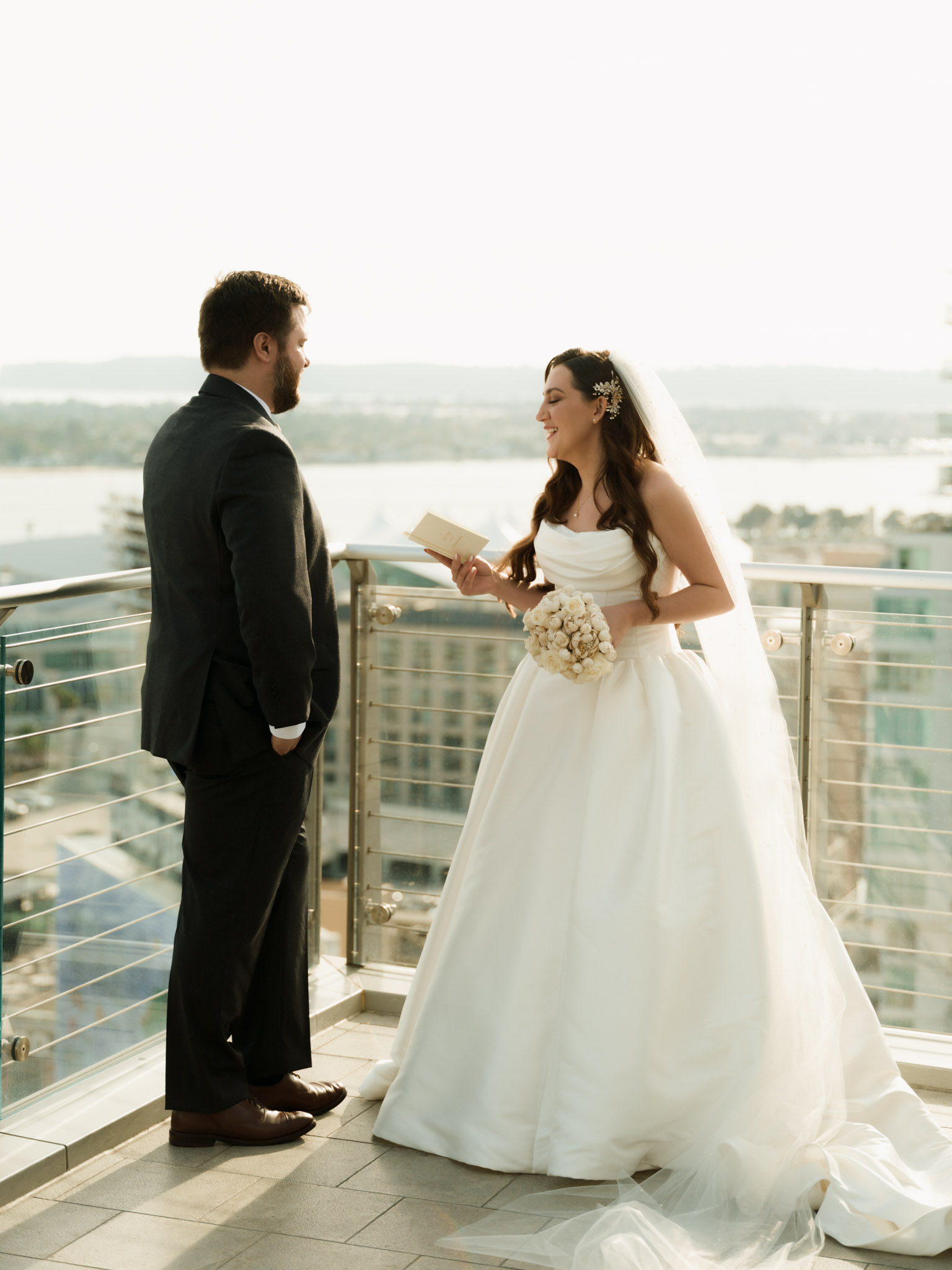 Bride in a strapless satin ballgown reading handwritten vows to the groom on an open-air rooftop terrace overlooking San Diego Bay.