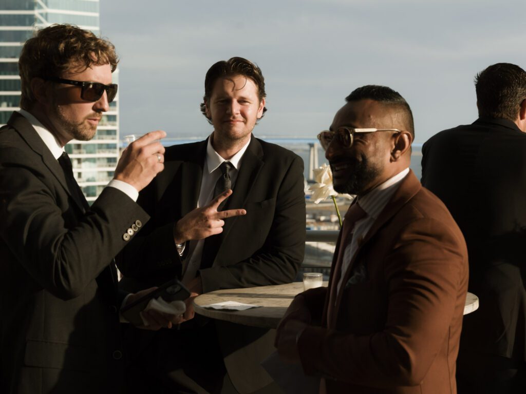 Well-dressed wedding guests wearing sunglasses conversing and laughing around a high-top cocktail table on an open-air terrace .