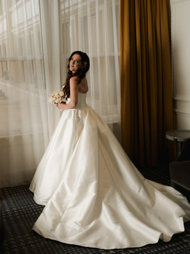 Full-length bridal portrait of the bride holding a white rose bouquet and glancing over her shoulder, displaying the buttons and train of her satin dress.