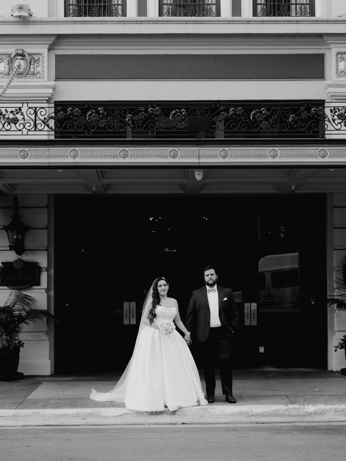 Black and white wide portrait of the bride in a satin ballgown and groom in a dark suit standing under the ornate wrought-iron marquee outside The US Grant Hotel.