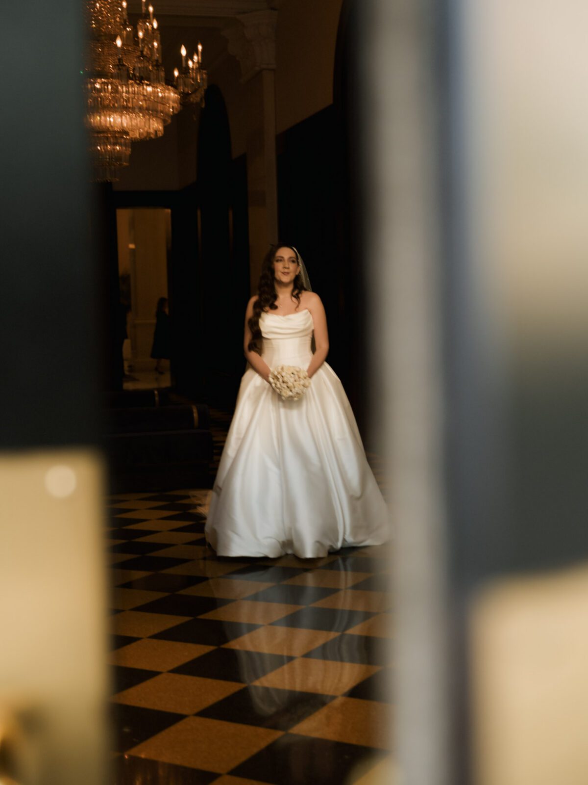 Bride in a strapless satin ballgown holding a white rose bouquet standing on the black and white checkered marble floor beneath a grand crystal chandelier.