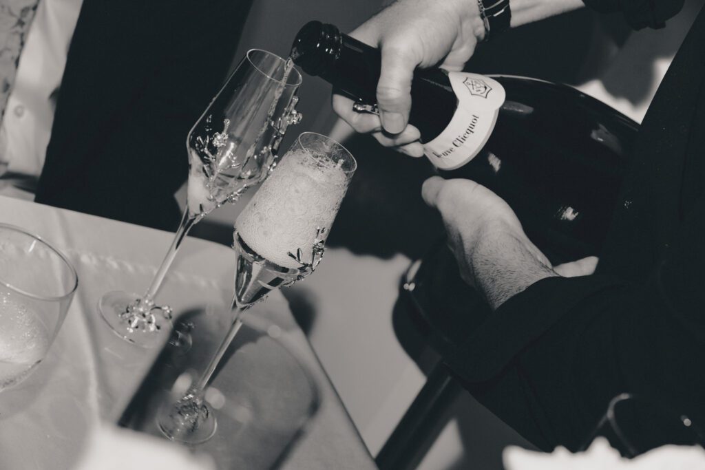 Black and white close-up of a hand pouring chilled Veuve Clicquot champagne into crystal flutes embellished with silver floral stems.