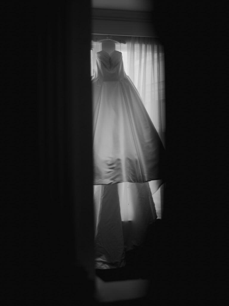Moody black and white photograph of a full satin wedding dress hanging in front of a sheer curtained hotel window.
