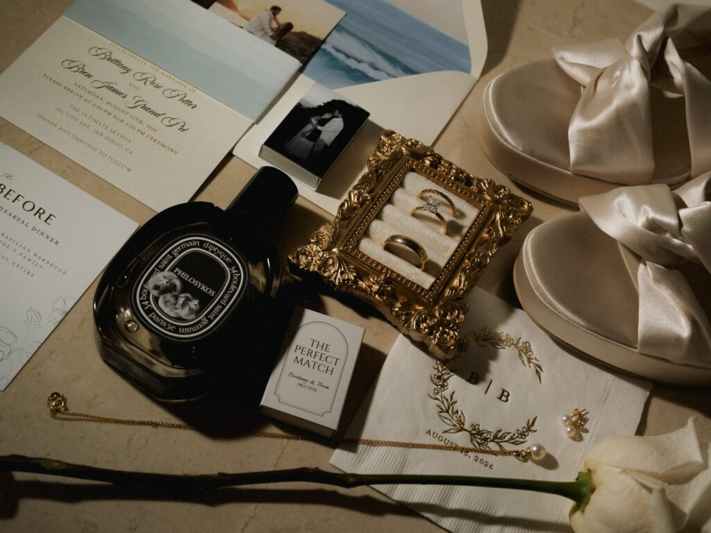 Elegant wedding flat lay with invitation suite, Diptyque Philosykos perfume, wedding rings in a mini gold frame, bridal platform heels, and pearl jewelry.