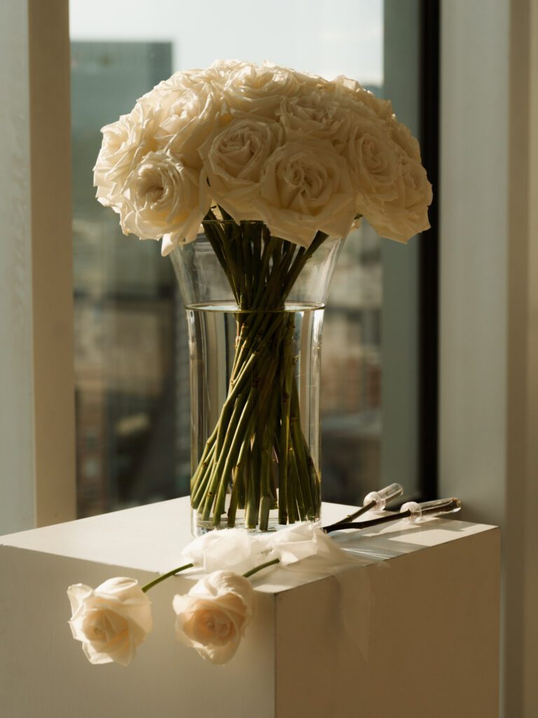 Lush round arrangement of reflexed white roses in a clear glass cylinder vase set on a modern white ceremony pedestal by a sunlit window .